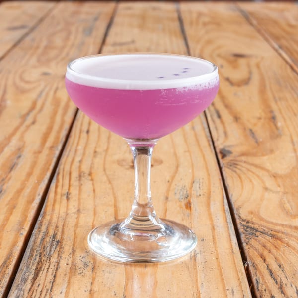 Vibrant pink cocktail with a white foam top and small dark specks, on a rustic wooden table.