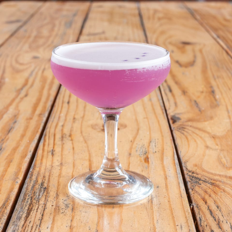 Vibrant pink cocktail with a white foam top and small dark specks, on a rustic wooden table.
