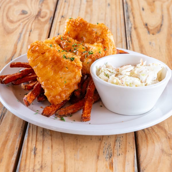 Golden-brown fish and chips with coleslaw on a white plate, resting on a rustic wooden table.