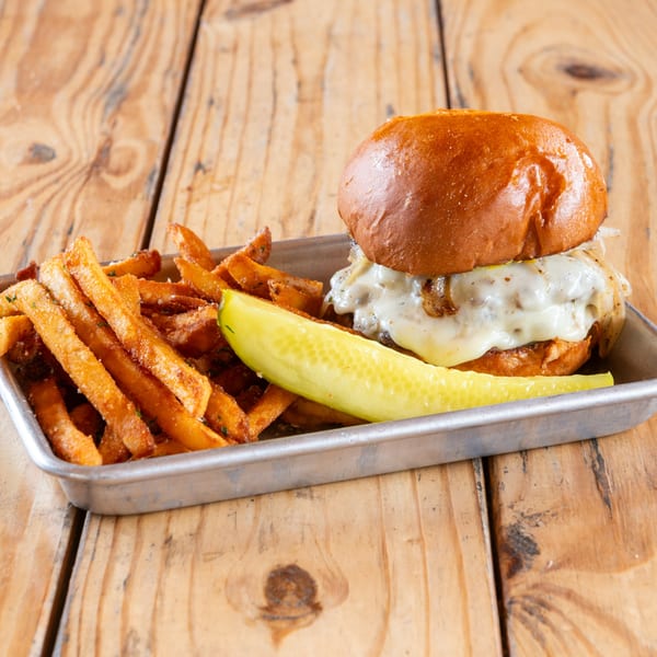 A burger with melted cheese and caramelized onions, sweet potato fries, and a pickle spear on a metal tray.
